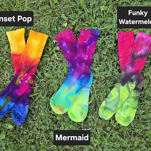 Peut inclure: Trois paires de chaussettes tie-dye. La premi&egrave;re paire, &eacute;tiquet&eacute;e Sunset Pop, pr&eacute;sente du jaune, de l'orange, du rose et du bleu. La deuxi&egrave;me, Mermaid, montre du violet, du bleu et du vert. La troisi&egrave;me, Funky Watermelon, affiche du rose, du noir et du vert lime.