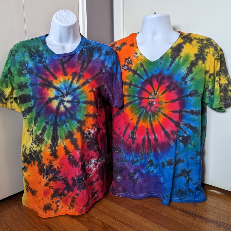 May include: Two tie-dye t-shirts on white mannequins. One is a crew neck, the other a V-neck. Both shirts feature a spiral pattern in a rainbow of colours, including red, orange, yellow, green, blue, and purple, with black accents.