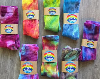 May include: Eight pairs of ribbed crew socks featuring various tie-dye color patterns including pink, orange, yellow, green, blue, and purple. Each pair is secured with a tan paper band displaying a rainbow-colored circular logo with the text Bree Happy Designs.
