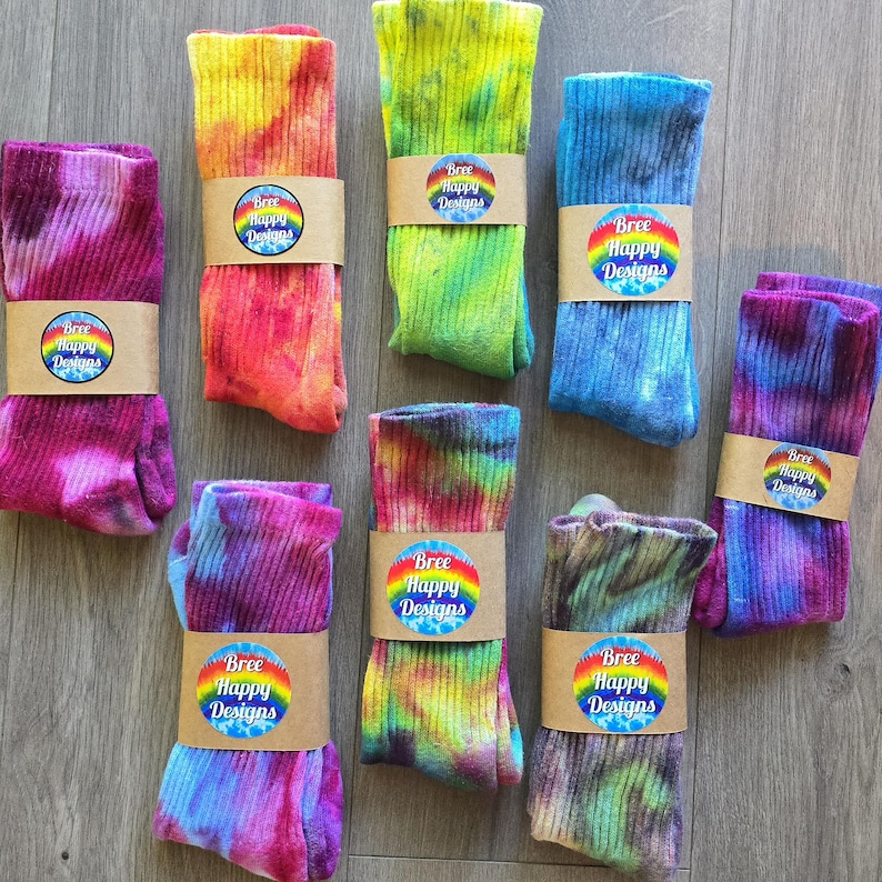 Peut inclure: Huit paires de chaussettes montantes c&ocirc;tel&eacute;es pr&eacute;sentant divers motifs tie-dye aux couleurs rose, orange, jaune, vert, bleu et violet. Chaque paire est maintenue par une bande de papier beige avec un logo circulaire arc-en-ciel et le texte Bree Happy Designs.