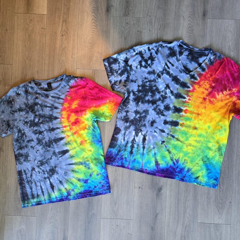 May include: Two tie-dye t-shirts. One is a crew neck, the other a V-neck. Both shirts feature a spiral pattern with a gradient of colours. The left side of each shirt is dark grey, while the right side transitions from red to yellow to blue.