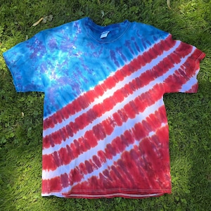 May include: A tie-dye t-shirt with a blue and purple upper section and red and white diagonal stripes. The shirt is made of soft material and has a crew neck. The shirt is laying on a bed of green grass.