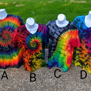 May include: Four tie-dye t-shirts on white mannequins. The shirts display spiral and swirled patterns in vibrant colours including red, orange, yellow, green, blue, and purple. Two shirts have crew necks, and two have V-necks. The shirts are labelled A, B, C, and D.