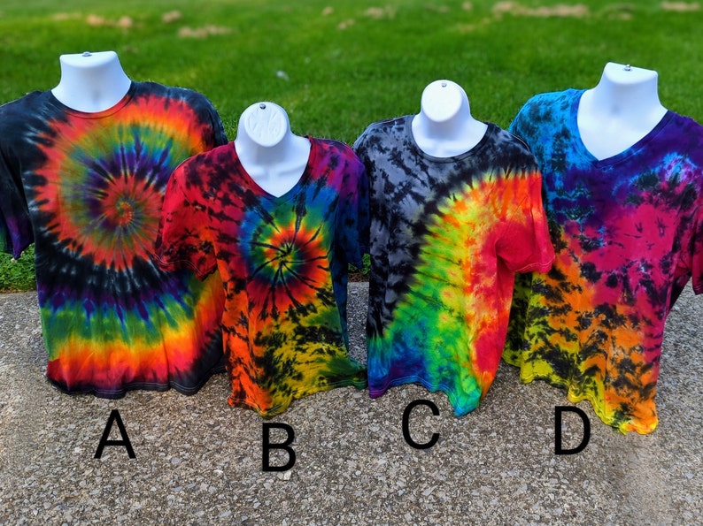 May include: Four tie-dye t-shirts on white mannequins. The shirts display spiral and swirled patterns in vibrant colours including red, orange, yellow, green, blue, and purple. Two shirts have crew necks, and two have V-necks. The shirts are labelled A, B, C, and D.
