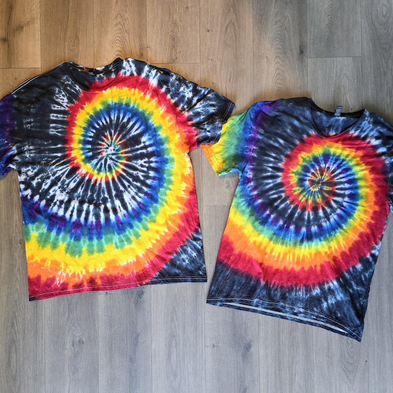 May include: Two short-sleeved tie-dye t-shirts. Each shirt has a spiral design with a blend of colours including red, orange, yellow, green, blue, and purple. The shirts have a black base colour.