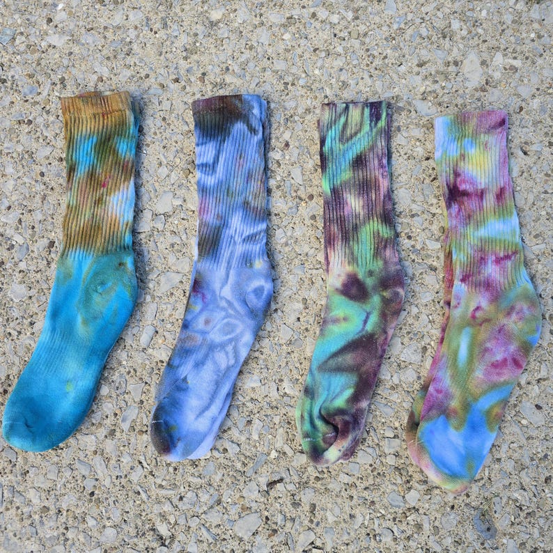 Peut inclure: Quatre chaussettes montantes avec des motifs tie-dye. La premi&egrave;re chaussette pr&eacute;sente des tons marron et bleu. La deuxi&egrave;me affiche des nuances de bleu et de gris. La troisi&egrave;me montre des teintes vertes, violettes et bleues. La quatri&egrave;me est rose, jaune et bleue.