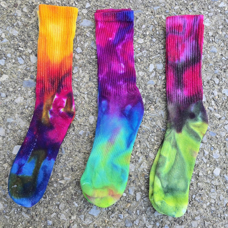 Peut inclure: Trois chaussettes mi-mollet avec des motifs tie-dye vibrants. La chaussette de gauche pr&eacute;sente des tons jaune, orange et magenta. Celle du centre affiche du magenta, du violet et du bleu. La droite combine magenta, noir et vert citron.