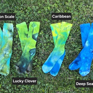 Peut inclure: Quatre paires de chaussettes montantes tie-dye dispos&eacute;es en rang&eacute;e. La premi&egrave;re paire est &eacute;tiquet&eacute;e Dragon Scale avec des tons verts et jaunes. La deuxi&egrave;me, Lucky Clover, pr&eacute;sente des motifs vert fonc&eacute;. La troisi&egrave;me, Caribbean, montre du jaune et du bleu, et la quatri&egrave;me, Deep Sea, affiche des nuances de bleu.