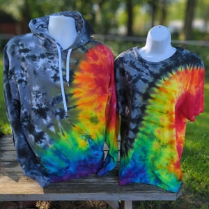 May include: Two tie-dye shirts on display. A hooded sweatshirt with a front pocket and a short-sleeved t-shirt. Both garments feature a spiral design with a colour gradient from black to rainbow hues, including red, orange, yellow, green, and blue.