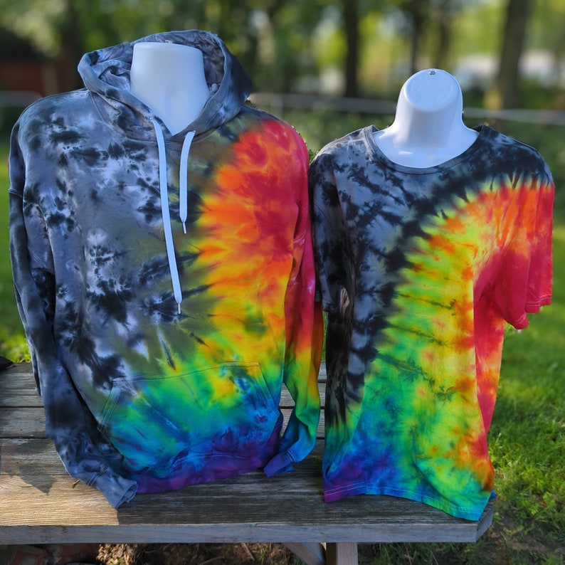 May include: Two tie-dye shirts on display. A hooded sweatshirt with a front pocket and a short-sleeved t-shirt. Both garments feature a spiral design with a colour gradient from black to rainbow hues, including red, orange, yellow, green, and blue.