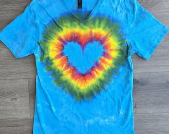 May include: A short-sleeve V-neck t-shirt in a bright sky blue color. The center of the shirt features a large, vibrant tie-dye heart pattern with concentric rings of yellow, orange, red, and purple, surrounded by a dark green border.