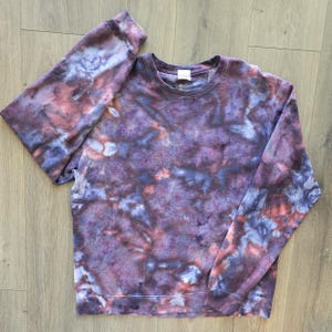 May include: A tie-dye crewneck sweatshirt in shades of purple, blue, and red. The long-sleeved top has a classic fit and a soft, textured appearance. The dye pattern creates a unique, marbled effect across the fabric.