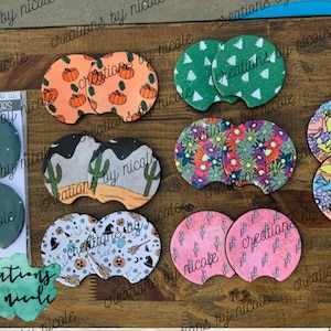May include: A set of 10 colorful, round, fabric-covered coasters with various designs. The designs include pumpkins, cacti, flowers, and a witch's hat. The coasters are labeled "Creations by Nicole".