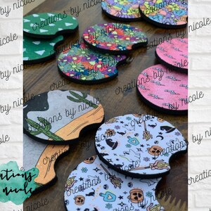 May include: A collection of colorful and patterned beverage coasters. The coasters are made of a soft, absorbent material and feature various designs, including cacti, flowers, and Halloween-themed patterns. The coasters are all round and have a slightly raised edge.