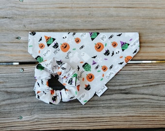 Könnte beinhalten: Ein weißes Stoff-Hundehalstuch mit Halloween-Motiv. Das Halstuch zeigt verschiedene gruselige Figuren, darunter Geister, Kürbisse, Fledermäuse und Hexen. Das Halstuch hat den Text "Boo!" und "Meow!" aufgedruckt. Das Halstuch wird mit einem weißen Stoff-Haargummi mit ähnlichem Halloween-Motiv geliefert.