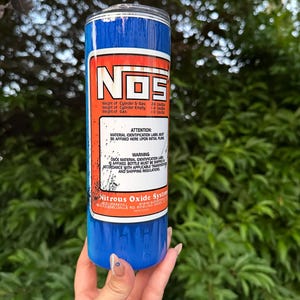 Pu&ograve; includere: Un bicchiere blu e bianco con la scritta "NOS" in grandi lettere bianche. Il bicchiere ha un'etichetta rossa e bianca con testo sul peso del cilindro e del gas. Il bicchiere &egrave; tenuto da una persona.