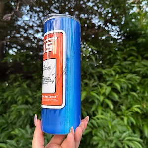 Pu&ograve; includere: Un bicchiere isolante blu con un'etichetta bianca e rossa. L'etichetta presenta il testo "TSE" e altre informazioni. Il bicchiere &egrave; tenuto contro uno sfondo di fogliame verde.