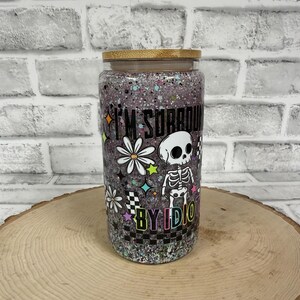 May include: A glass tumbler with a wooden lid, filled with glitter and a design that includes a skeleton, flowers, and the text "I'M SURROUNDED BY IDIOTS". The tumbler is on a wooden surface.