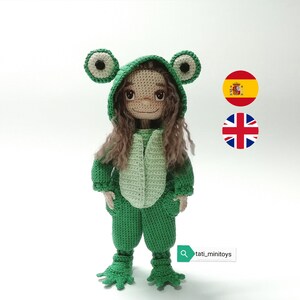 May include: A crocheted doll wearing a green frog costume with large eyes. The doll has brown hair and a sweet expression. The doll is standing on a white background.