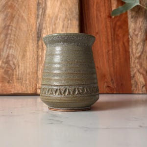 May include: A handmade ceramic vase with a textured, earthy green glaze. The vase features a decorative band of repeating triangular patterns near the base. The vase is approximately 10 cm tall and has a rustic, artisanal aesthetic.