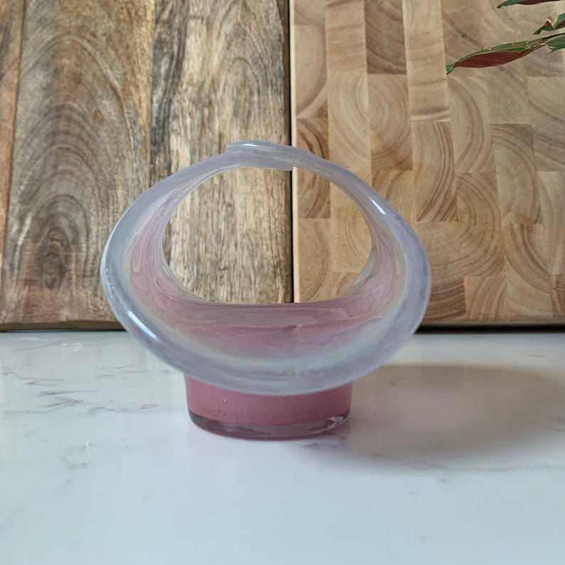 May include: A pink and white glass bowl with a unique curved design. The bowl is made of clear glass with a pink tint and has a white rim.