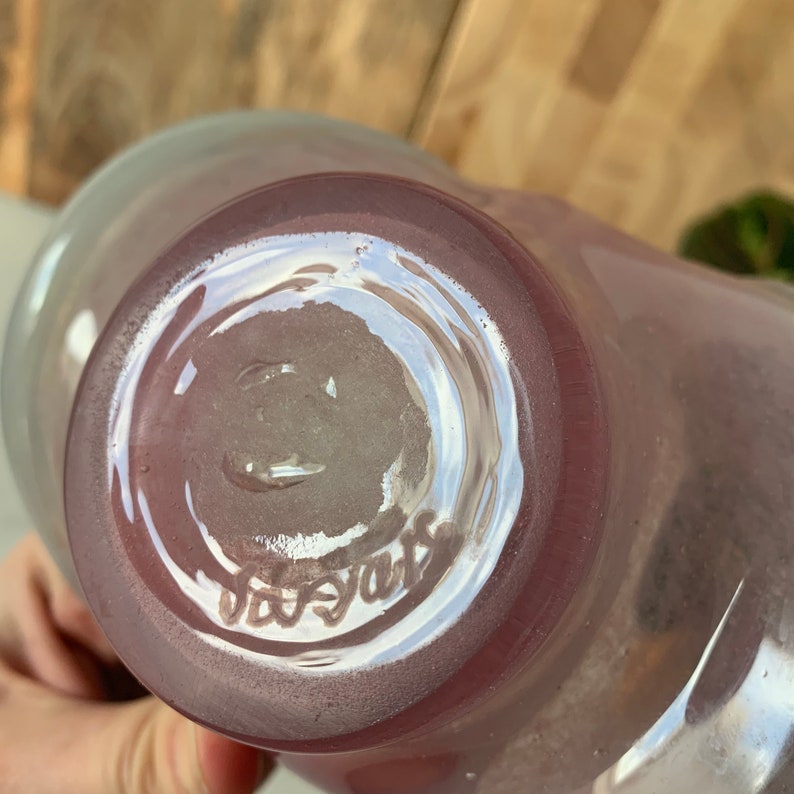 May include: The bottom of a clear glass vase with a pink tint. The vase has a circular base and the words "Legras" are etched into the bottom.