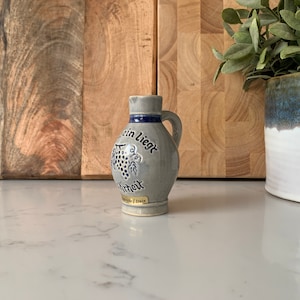 May include: A small grey ceramic jug with a blue and white design. The jug has a handle and a spout. The text "Der Wein Liegt Im Blut" is written on the jug.