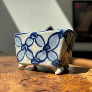 May include: A small, rectangular ceramic planter with four short feet. The off-white surface features a hand-painted, repeating blue floral pattern consisting of interconnected four-petaled blossoms with dark blue outlines and light blue centres.