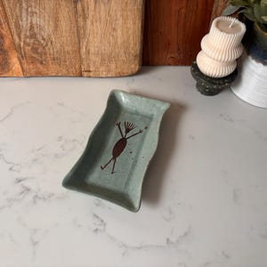 May include: A light blue ceramic dish with a wavy edge. The dish features a brown abstract figure design. The dish is speckled and appears handmade. The dish is approximately 15 cm long and 10 cm wide.