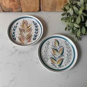 May include: Two ceramic plates with blue rims and white backgrounds. Each plate features a stylised floral design with yellow, orange, and brown accents.