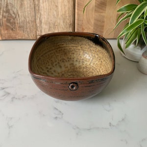 May include: A square ceramic bowl with a brown and tan speckled glaze. The bowl has a black rim and a small black knob on the side.