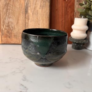 May include: A handcrafted ceramic bowl with a dark green and black glaze. The bowl has a rounded shape with a slightly flared rim. The interior is a deep, glossy green, while the exterior has a mottled, textured appearance.
