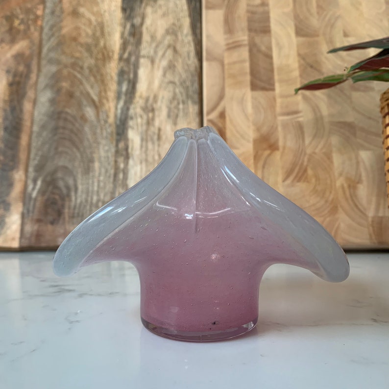 May include: A small, clear glass vase with a pink and white ombre effect. The vase has a unique, flower-like shape with four petals.
