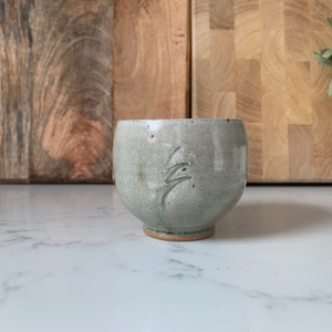 May include: A small, light green ceramic bowl with a carved design. The bowl has a slightly rounded bottom and a smooth, matte finish.