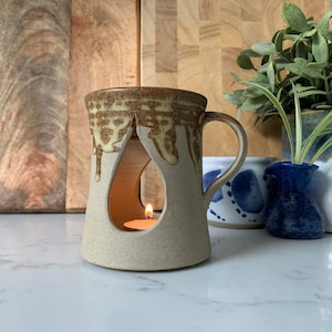 May include: A ceramic tea light holder with a brown and white glaze. The holder has a teardrop-shaped opening for a tea light and a handle. The holder is sitting on a white surface.