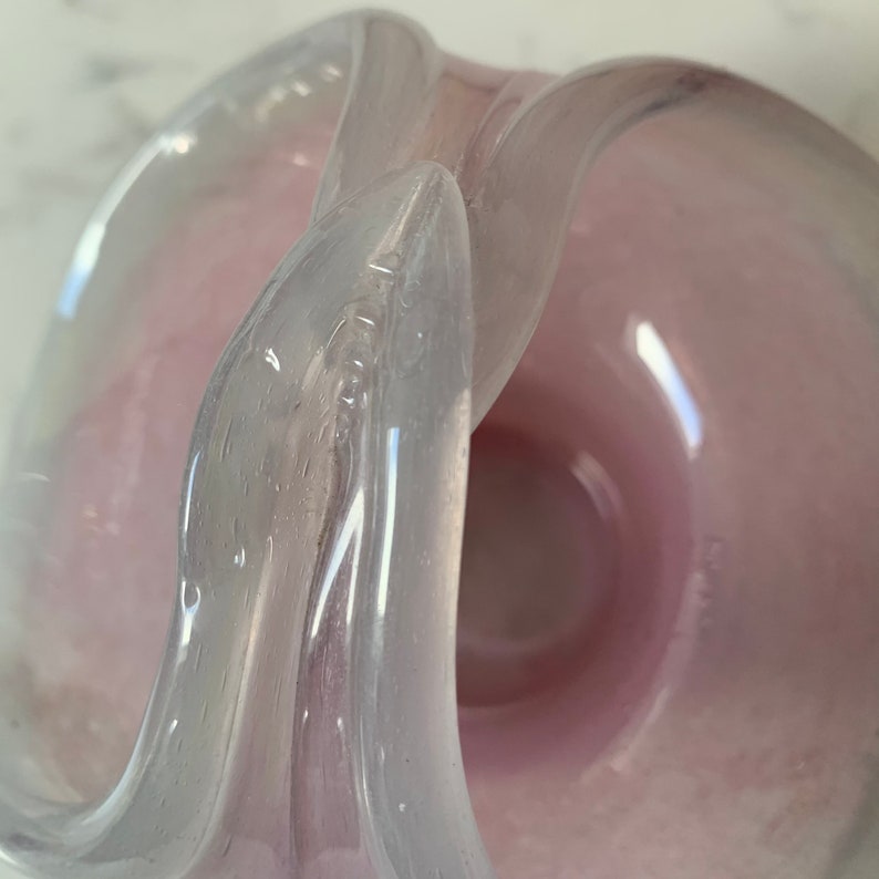 May include: A pink and white glass bowl with a unique, twisted design. The bowl is made of clear glass with a pink tint and has a smooth, glossy finish.
