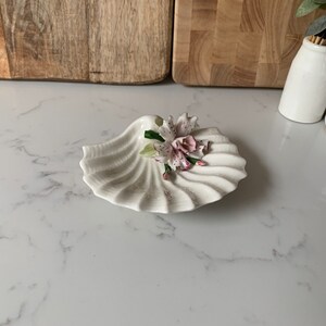 May include: A white porcelain seashell dish with a delicate pink and white flower accent. The dish is ideal for holding small trinkets or jewellery.