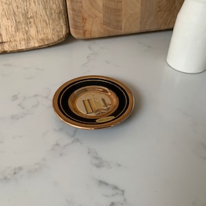 May include: A small, round, gold-rimmed plate with a black border. The plate features a depiction of the New York City skyline with the World Trade Center Towers in the centre. The plate is inscribed with "New York" and "24kt Gold."