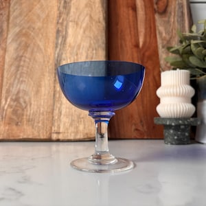 May include: A vibrant cobalt blue coupe glass with a clear stem and base. The glass features a wide, shallow bowl and a classic design. The stem is slender, and the base is round. The glass is set against a wooden background.