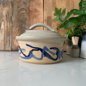 May include: A ceramic casserole dish with a light grey glaze and blue and white abstract design. The dish has a lid with a handle.