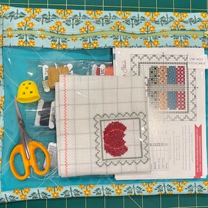 May include: A cross-stitch kit in a teal and floral patterned zippered pouch. The kit includes scissors, thread, a thimble, and a cross-stitch pattern with a red apple design. The pouch has a clear front panel for visibility.