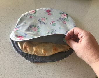 May include: A gray and floral fabric bread cover with a round shape. The cover is being lifted to reveal a round, flat bread inside.