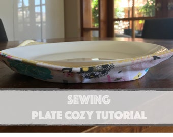 May include: A close-up of a plate cozy made from fabric with a floral pattern. The cozy is folded around a white plate and has a handle on each side. The text "Sewing Plate Cozy Tutorial" is visible at the bottom of the image.