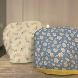 May include: Two fabric covers for kitchen appliances. One cover is white with a brown and white pattern of birds. The other cover is blue with a white daisy pattern and a yellow trim.