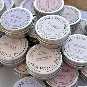 May include: A collection of round silver metal tins containing thread conditioner, each labelled The Stitch Wax by The Craftaholic Club. The labels feature various scent names like Slow Sunday, Soft Focus, and Golden Hour, with a net weight of 14g.