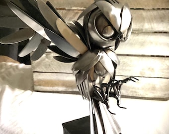 May include: A silver metal sculpture of an owl with outstretched wings. The owl is perched on a black metal base. The sculpture is made from recycled metal.