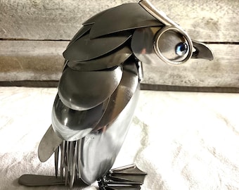 May include: A silver metal sculpture of a penguin made from spoons. The penguin is standing on a white surface. The penguin has blue eyes.
