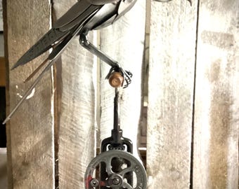 May include: A metal sculpture of a bird perched on a mechanical device. The bird is constructed from spoons and other metal pieces. The sculpture is mounted on a wooden base, creating a unique and industrial art piece.