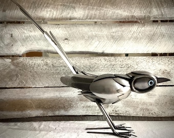 May include: A silver metal sculpture of a bird with a long tail. The bird is made from recycled materials and has a unique, whimsical design.