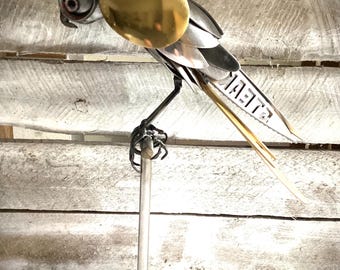May include: A metal sculpture of a bird with a gold wing and a silver body. The bird is perched on a metal stand.